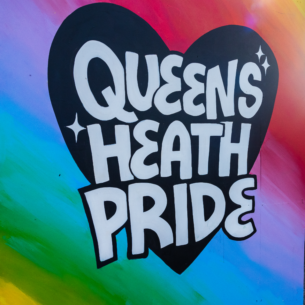 Hand painted sign. 
Rainbow background. 
Black Heart. 
White text reading: Queens Heath Pride